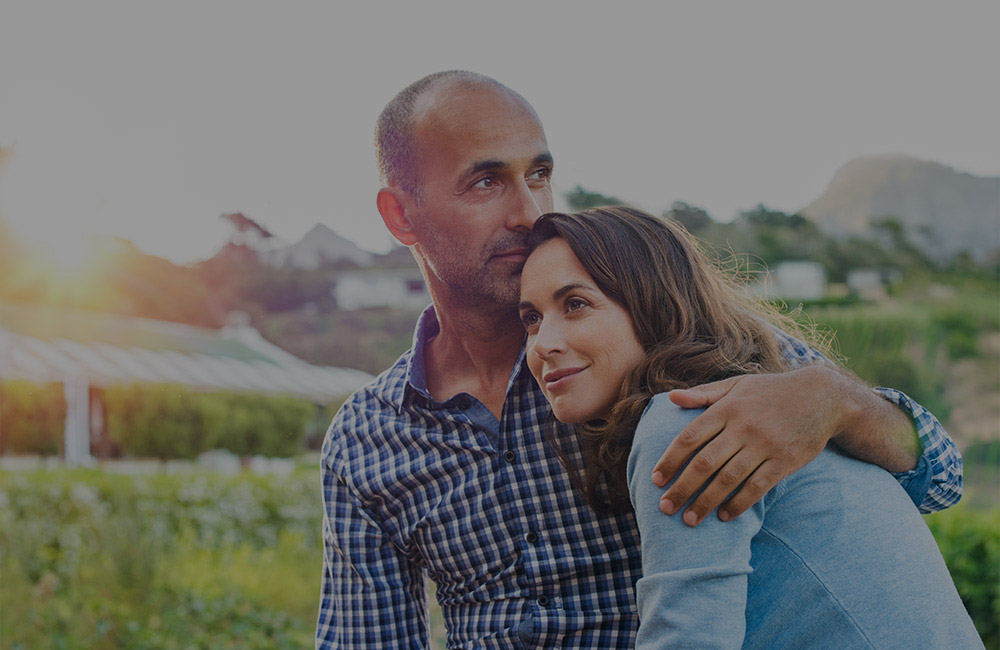 A man and woman - he has his arm wrapped around her shoulders as they look in different directions